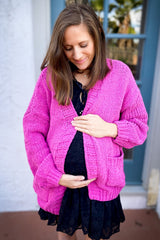 woman wearing a hot pink chunky knit maternity cardigan and a black dress, gently cradling her baby bump