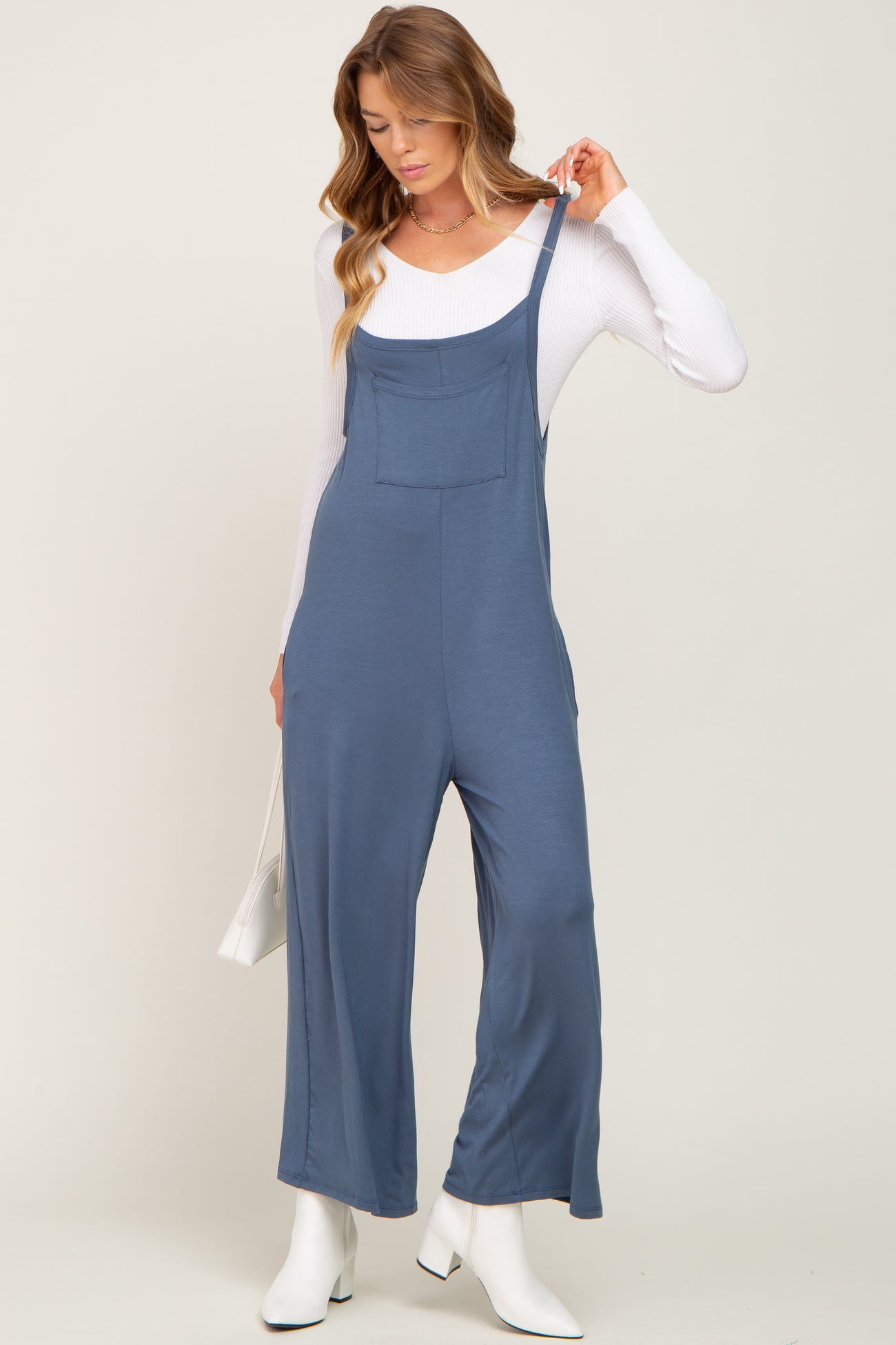 woman wearing a navy blue jumpsuit with adjustable straps and a front pocket, styled with a white long sleeve top and boots