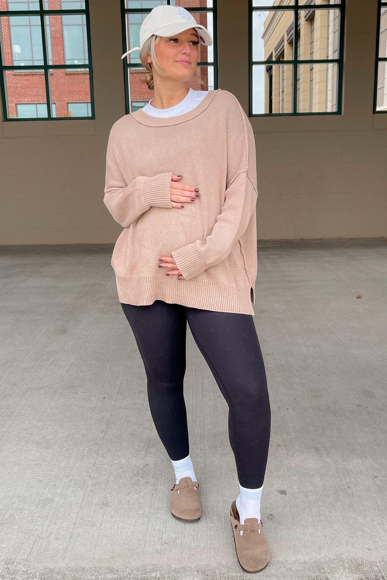 woman wearing a beige oversized maternity sweater and black leggings, holding her baby bump, styled with a baseball cap