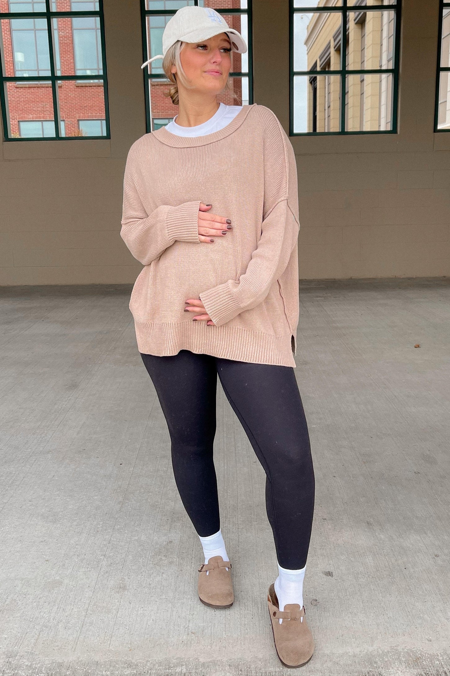 woman wearing a beige oversized maternity sweater and black leggings, holding her baby bump, styled with a baseball cap