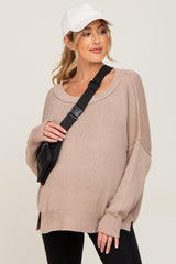 woman wearing a beige oversized maternity sweater with dropped shoulders and side slits, accessorized with a black crossbody bag and cap
