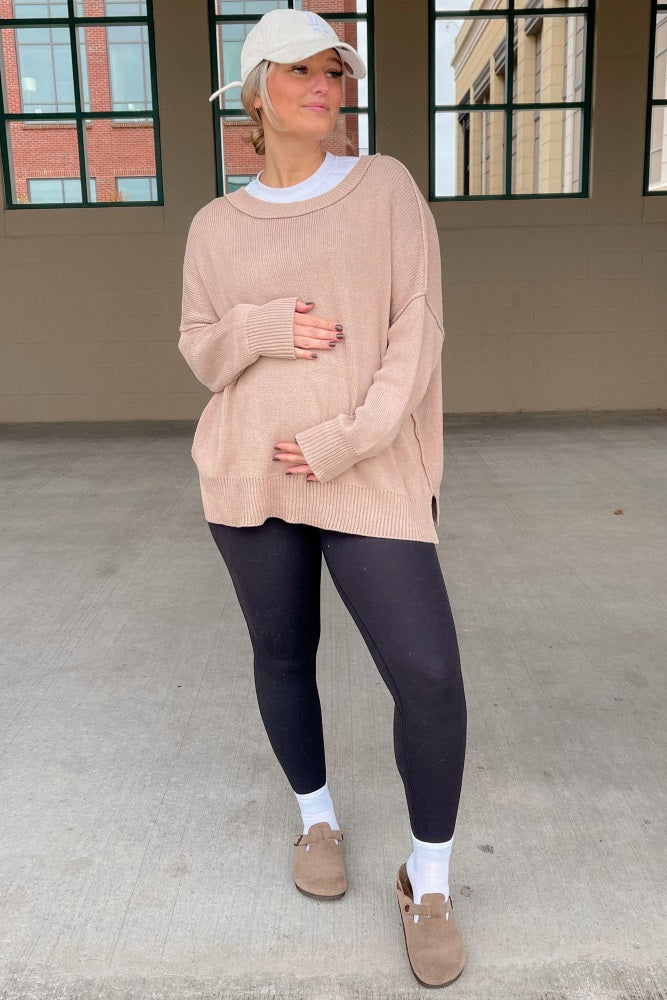 woman wearing a beige oversized maternity sweater and black leggings, holding her baby bump, styled with a baseball cap
