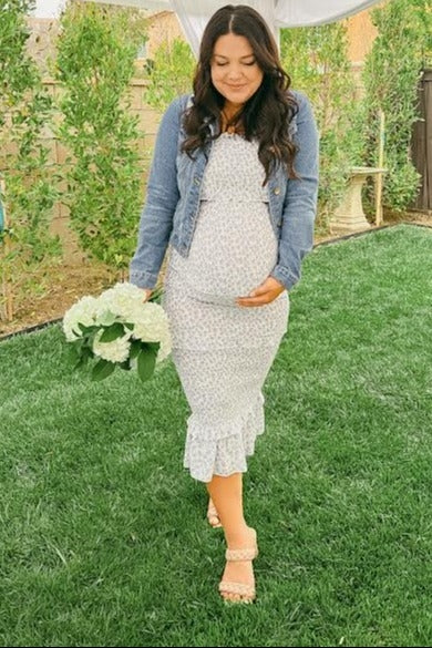 woman wearing a white floral maternity dress with ruffle hem and denim jacket, holding a bouquet and showing her baby bump