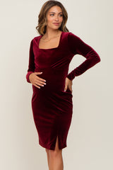 woman wearing a burgundy long sleeve velvet maternity dress, smiling and holding her baby bump