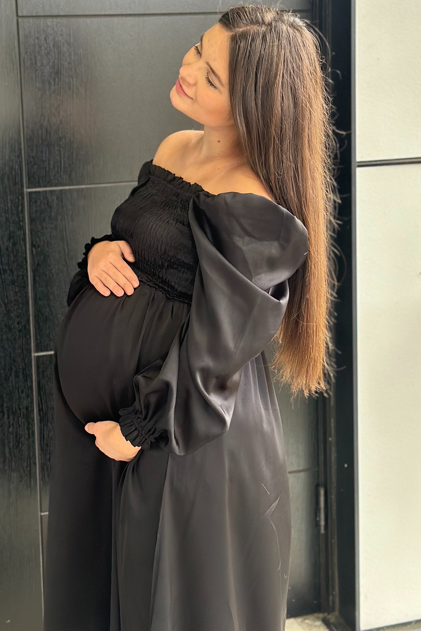 woman wearing a black off-shoulder maternity dress, showing her baby bump while posing outdoors