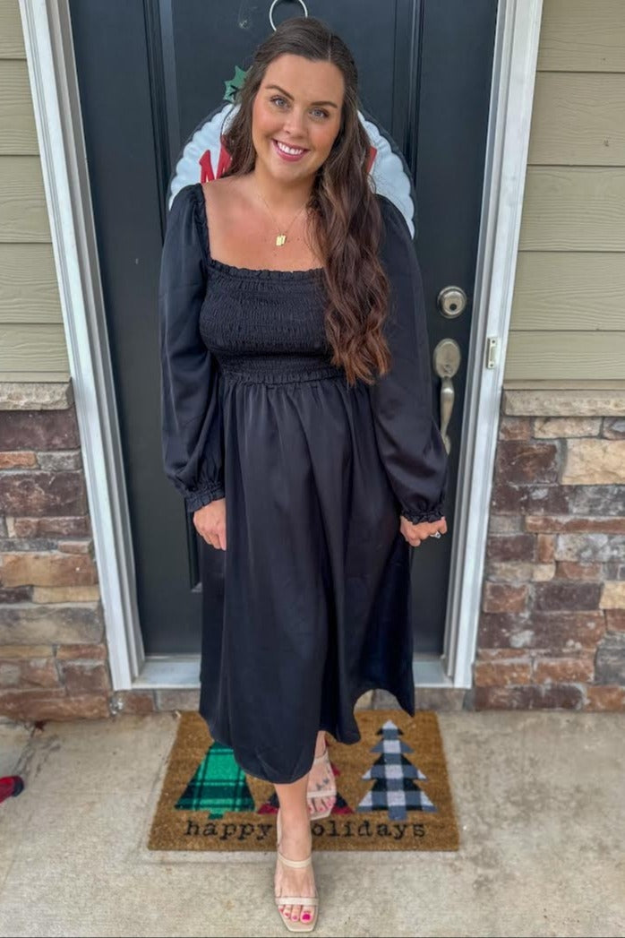 woman wearing a black maternity dress with puff sleeves and a smocked bodice, standing on a welcome mat