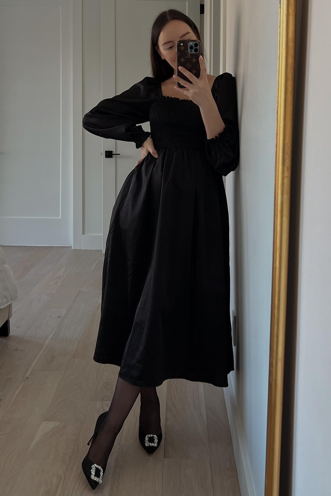 woman wearing a black smocked midi dress with long sleeves and high heels, posing in front of a mirror