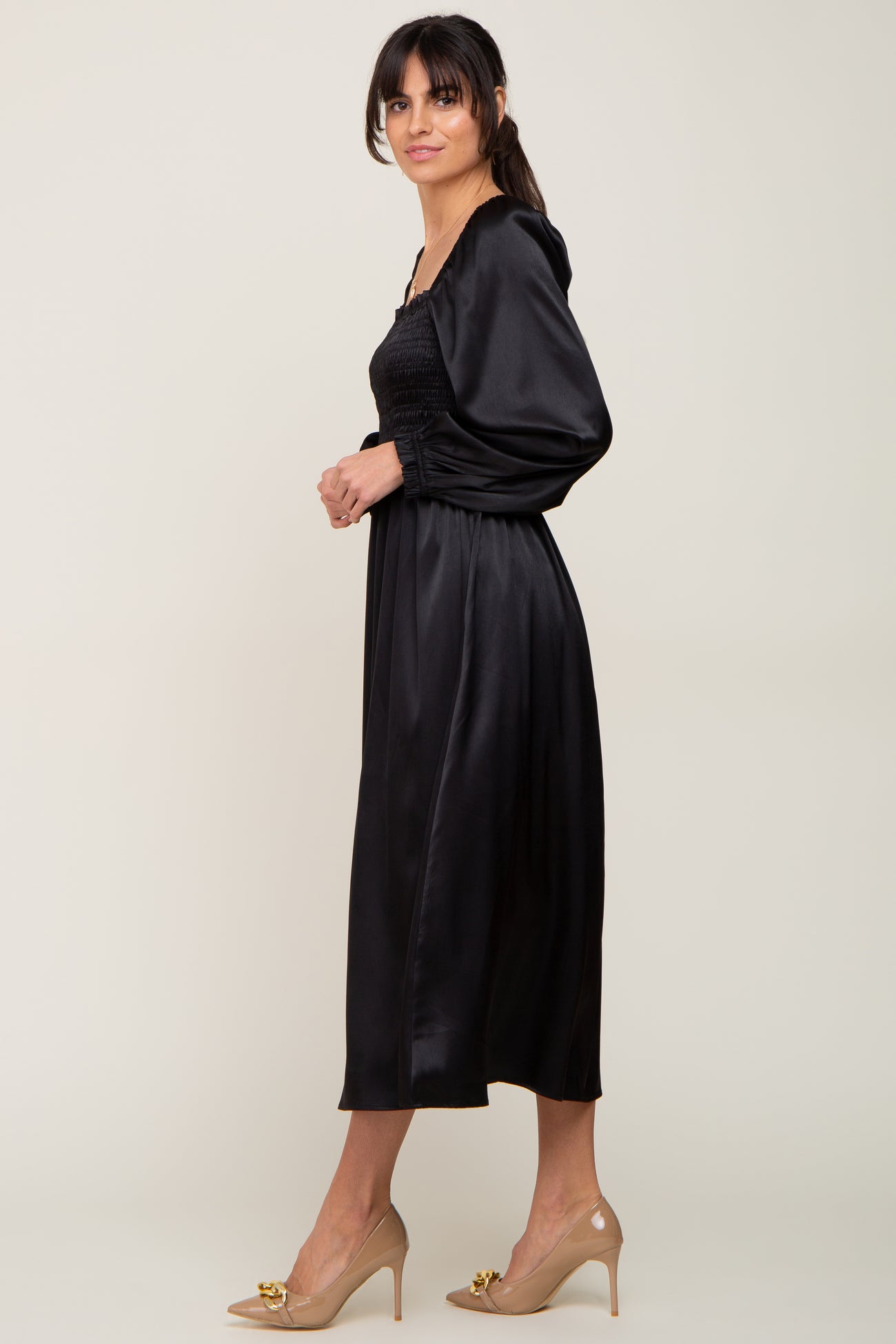 woman wearing a black satin long sleeve midi dress with a smocked bodice and flowing skirt, paired with nude heels