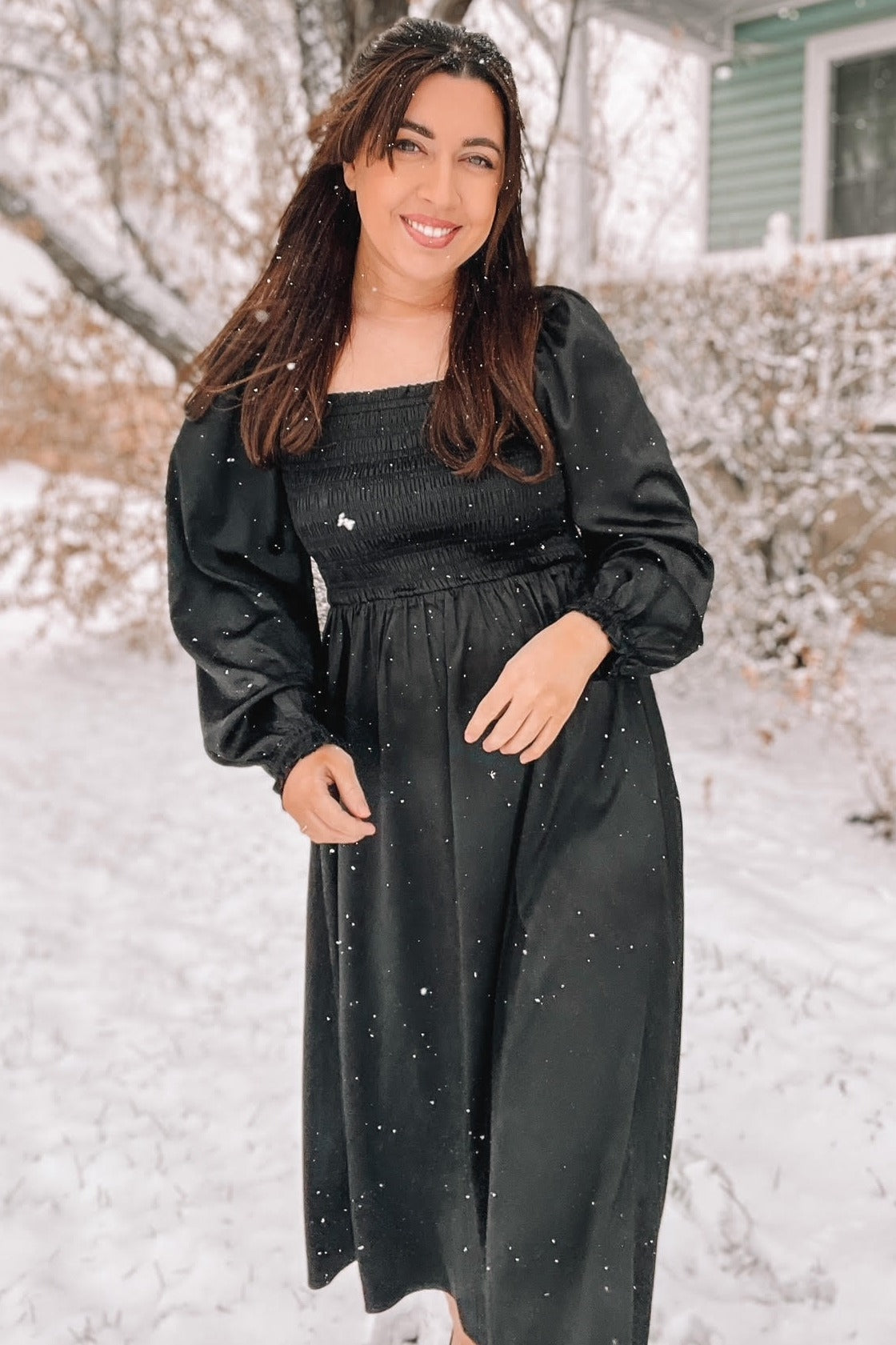 woman wearing a black smocked long sleeve dress, standing in the snow with a smiling expression