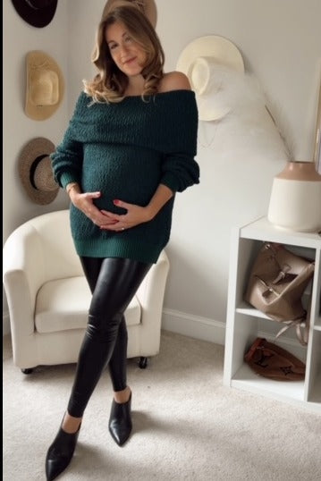 woman wearing a dark green off-shoulder maternity sweater and black leather leggings, holding her baby bump