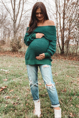 woman wearing a green off-the-shoulder maternity sweater and distressed jeans, holding her baby bump outdoors
