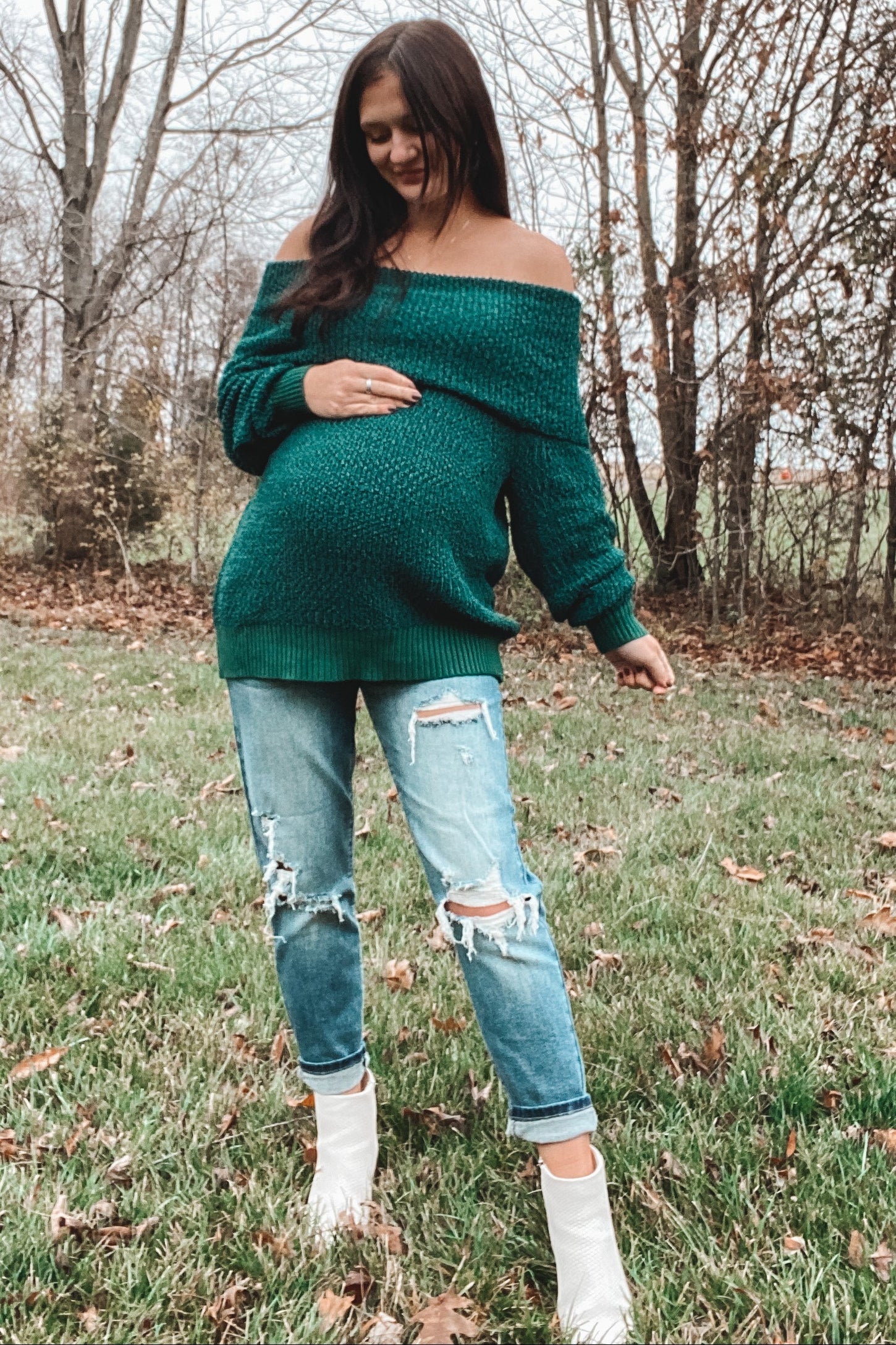 woman wearing an off-the-shoulder green maternity sweater and distressed jeans, showing her baby bump in a natural setting