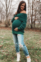 woman wearing a dark green off-the-shoulder maternity sweater and holding her baby bump, paired with distressed light blue jeans