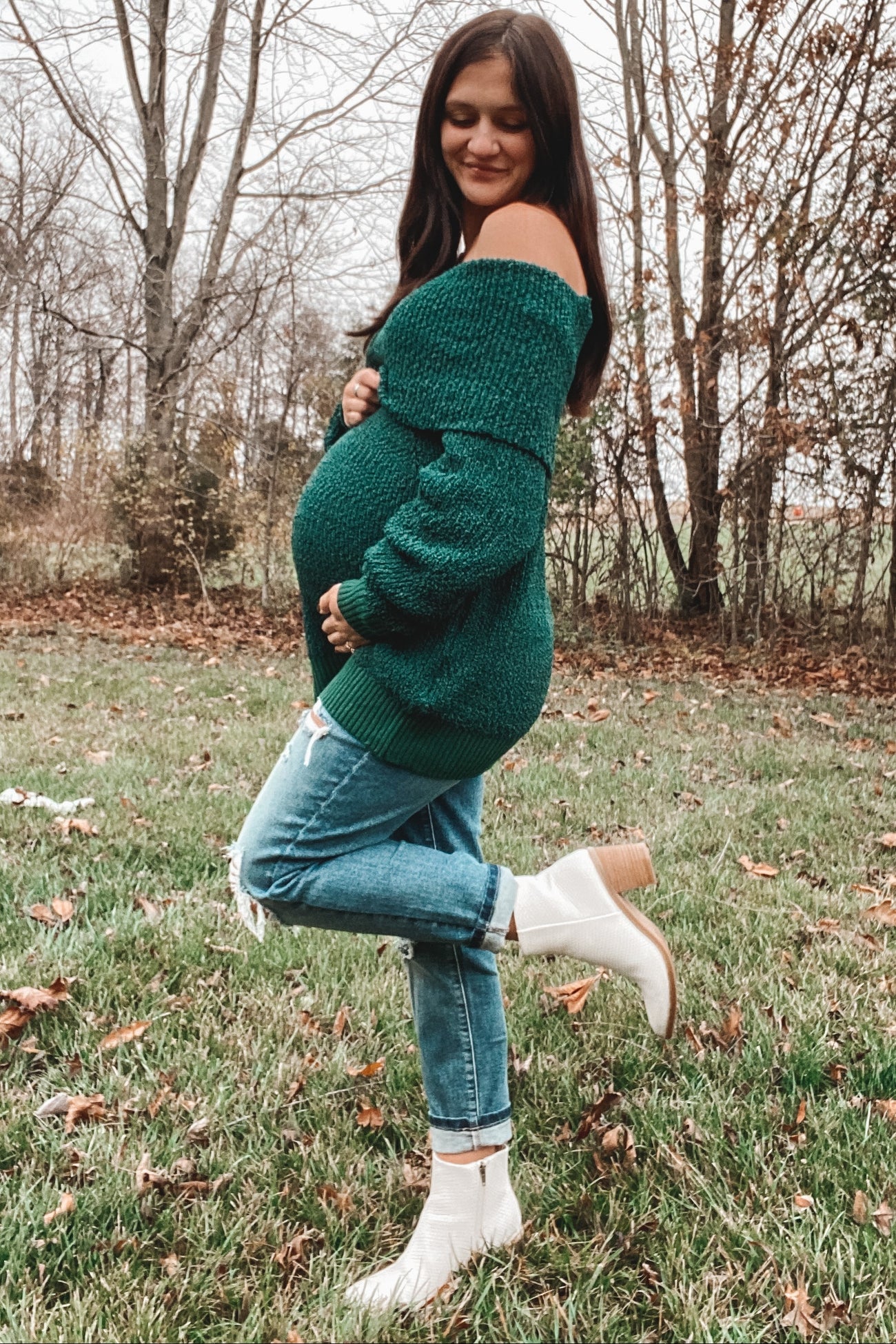 woman wearing a green off-shoulder maternity sweater and distressed jeans, showcasing her baby bump in a grassy outdoor setting
