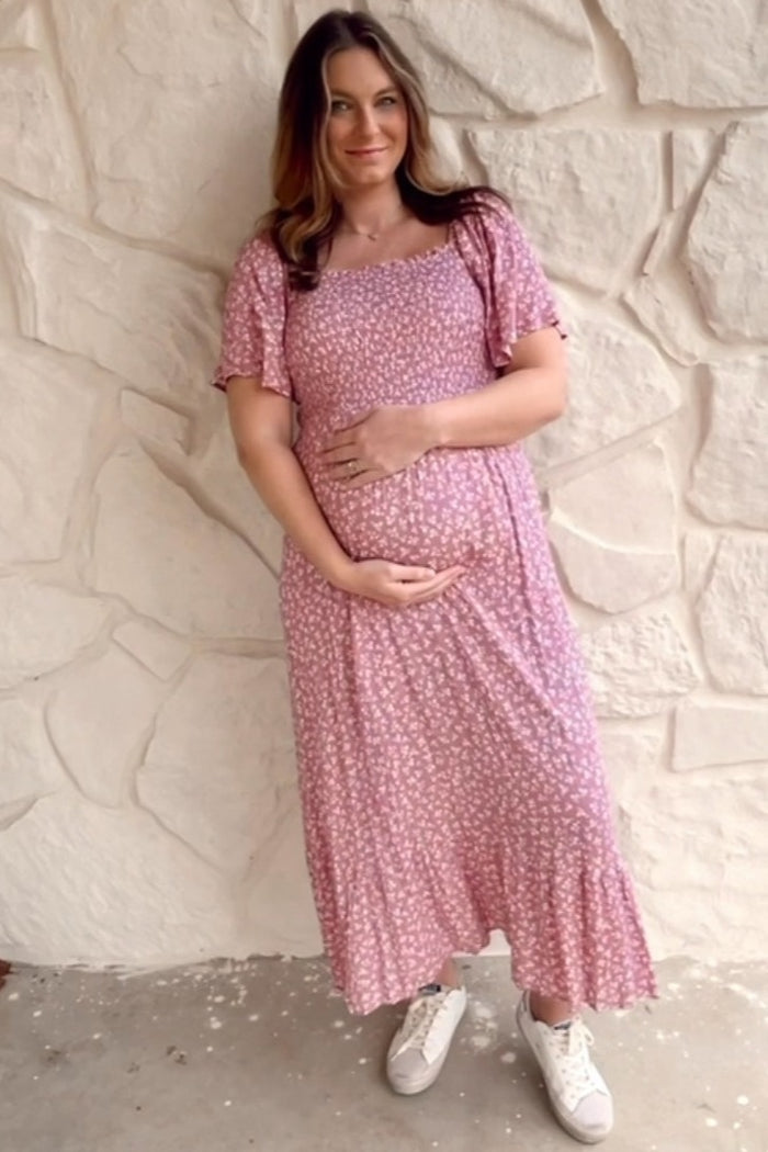 pregnant woman in pink floral maternity maxi dress holding her baby bump against a white stone wall