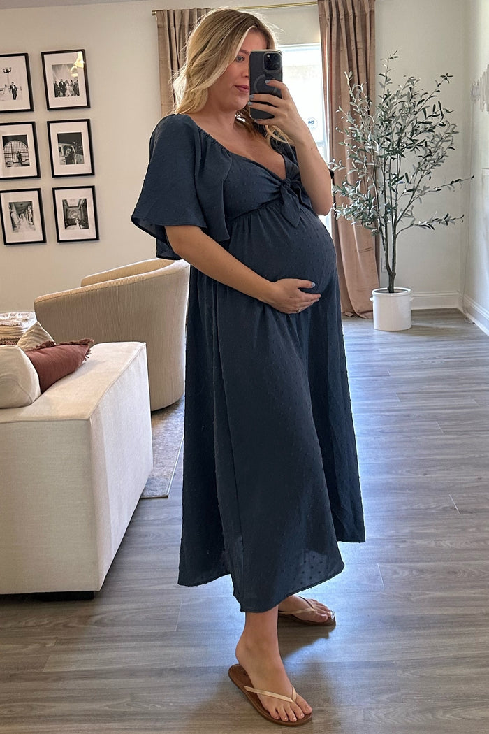 woman wearing a navy blue short sleeve maternity dress with a tied front, showing her baby bump while posing in a living room