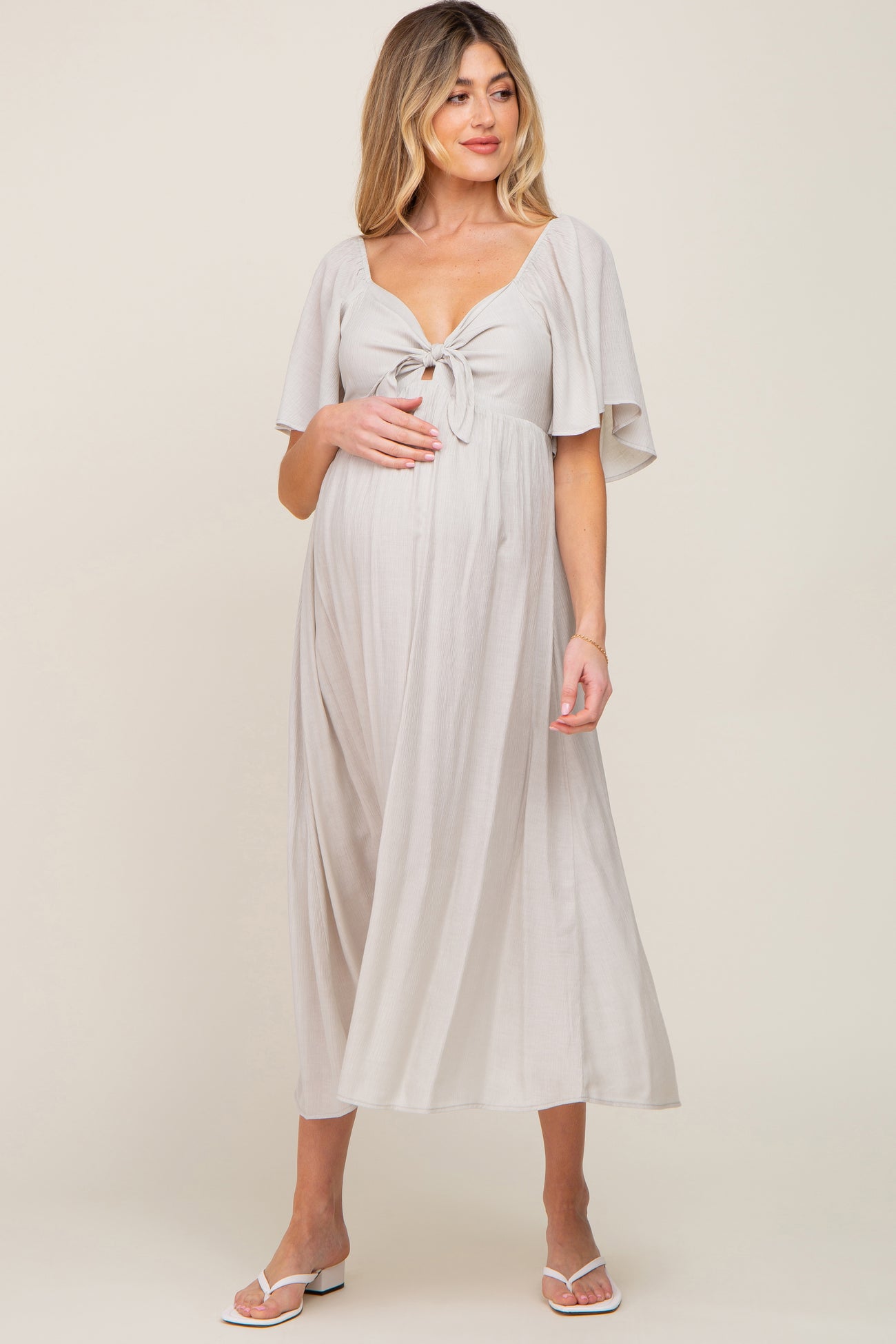 woman wearing a light gray maternity dress with a tied front and flutter sleeves, holding her baby bump
