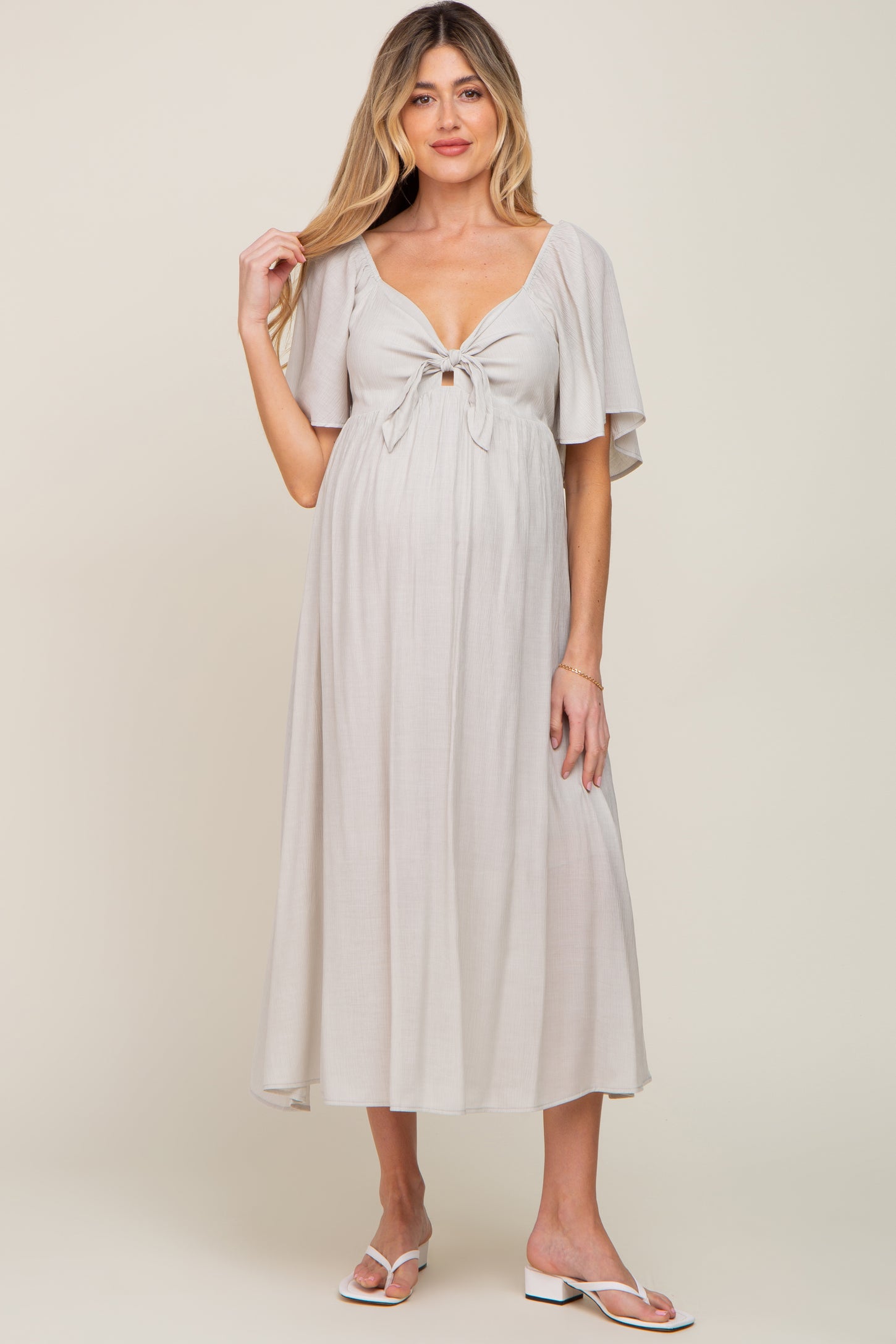 woman wearing a beige maternity dress with short flutter sleeves and a tie-front, showing her baby bump