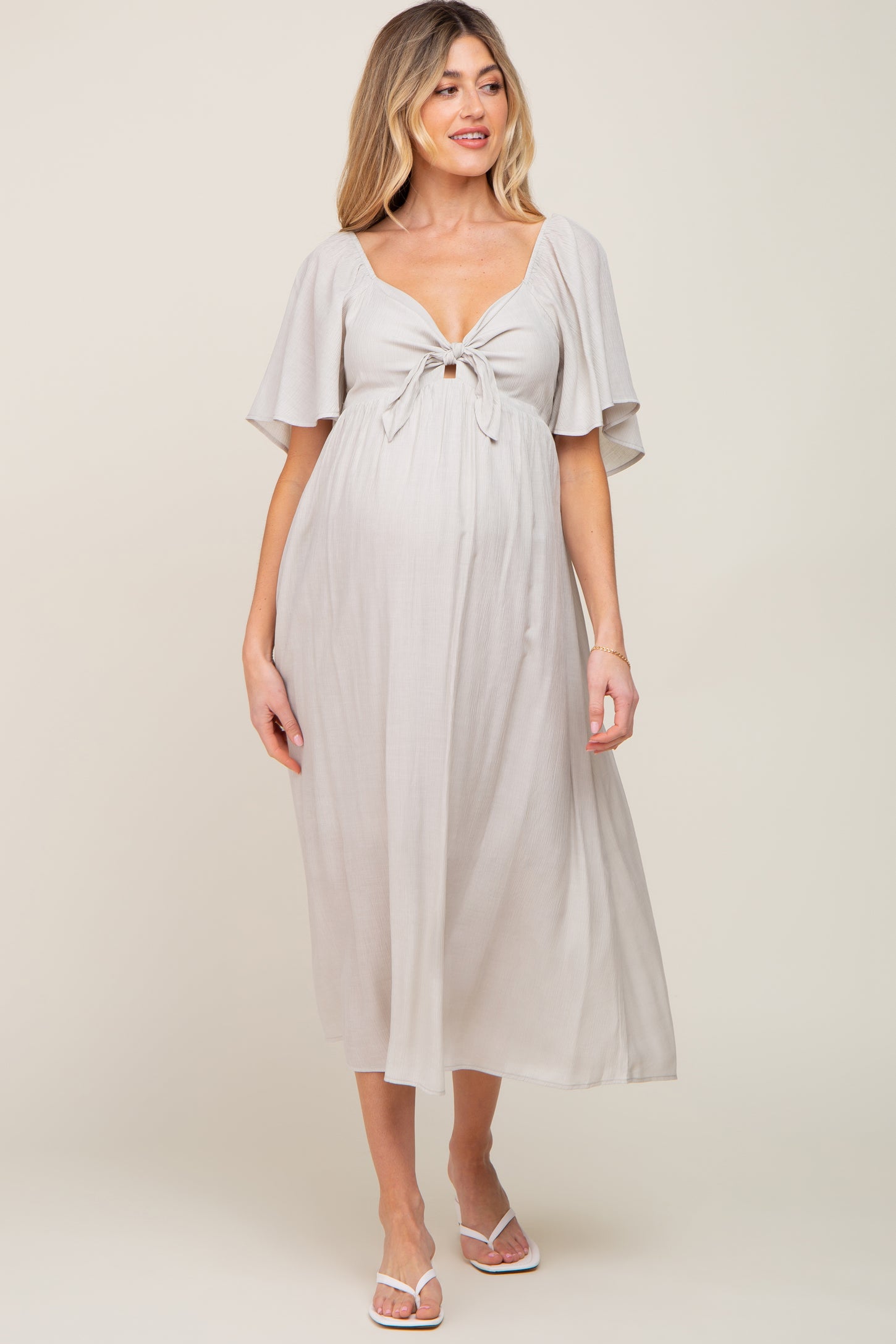 woman wearing a light gray maternity dress with a tie front and flutter sleeves, showcasing her baby bump