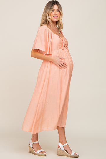 woman wearing a peach maternity dress with flutter sleeves and tie detail, gently holding her baby bump