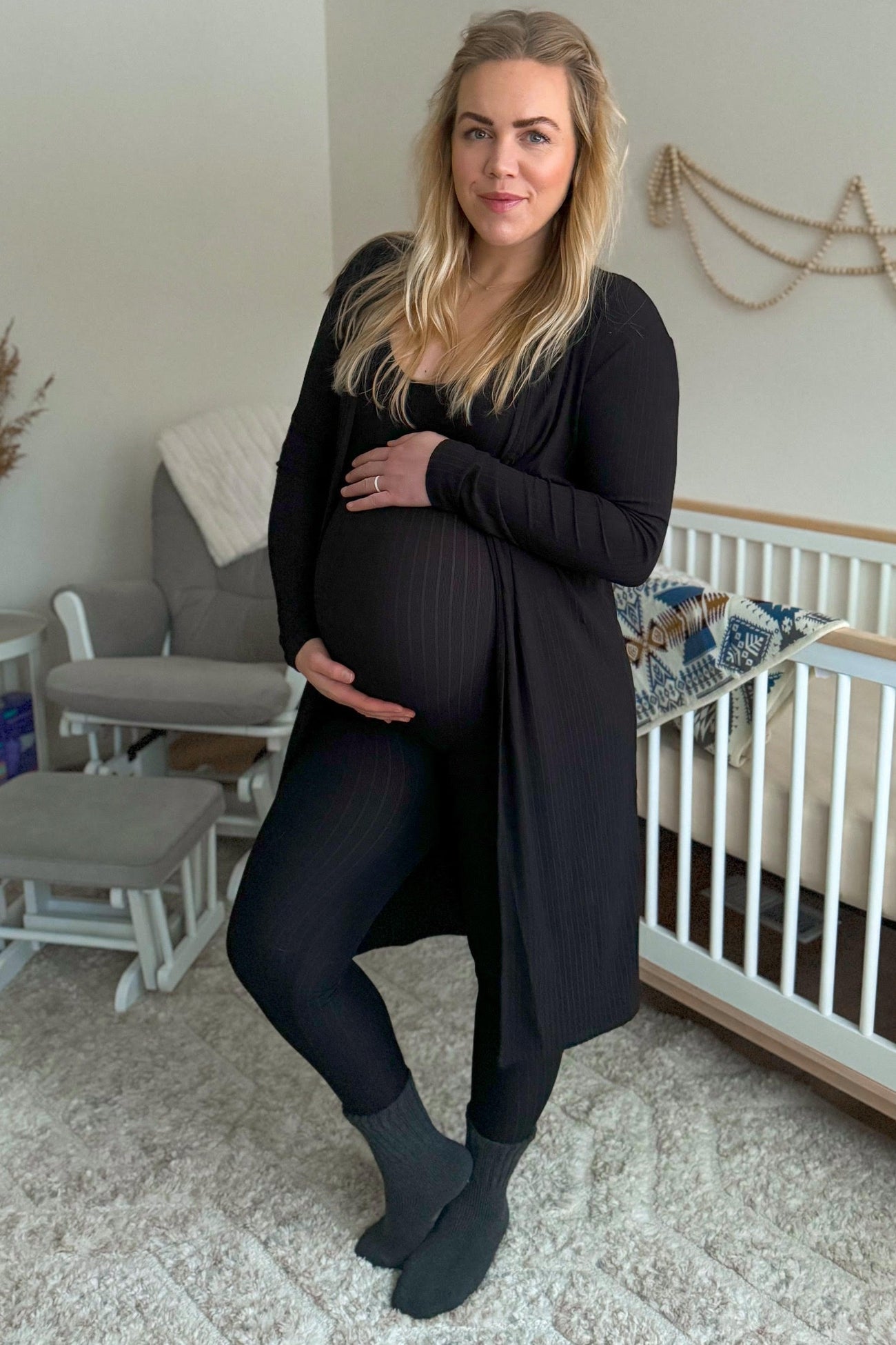 woman wearing a black maternity cardigan and leggings, holding her baby bump while standing in a nursery