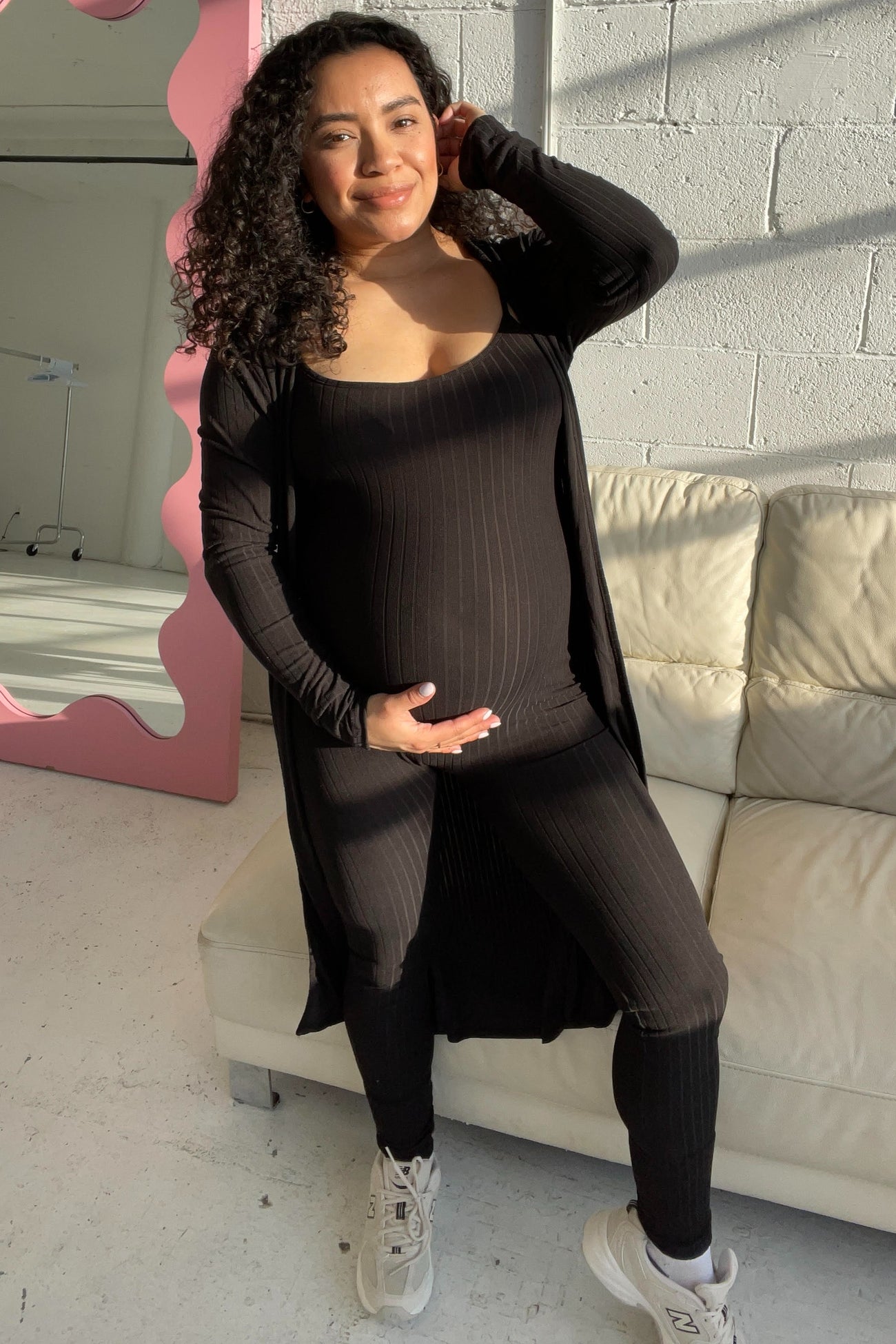 woman wearing a black ribbed maternity outfit with a long cardigan, showing her baby bump, standing in a bright room