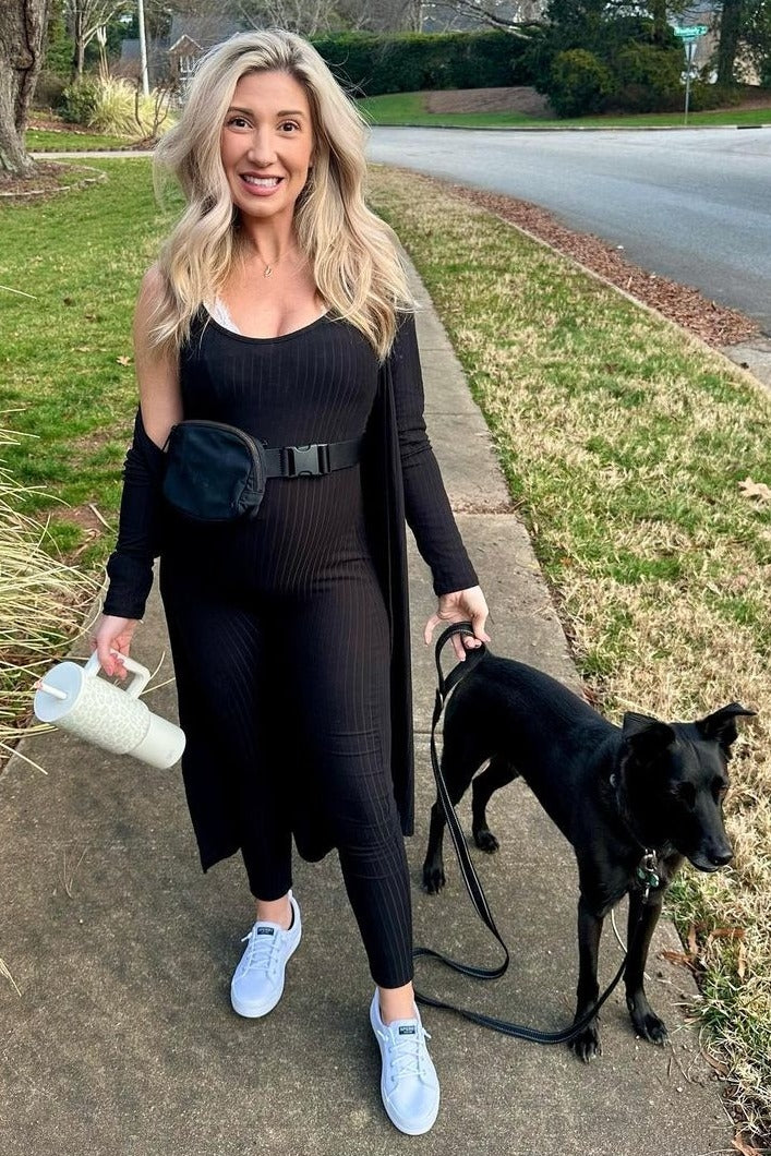 woman wearing a black ribbed maternity jumpsuit and a long black cardigan, walking a dog while holding a water bottle