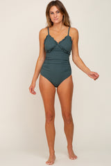 woman wearing a dark green ruffled one-piece swimsuit, standing barefoot against a neutral background