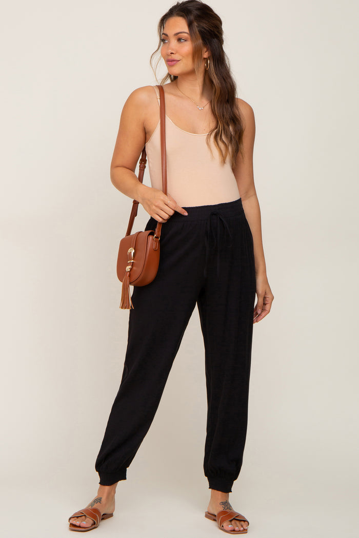 woman wearing black maternity joggers and a light beige tank top, carrying a brown crossbody bag