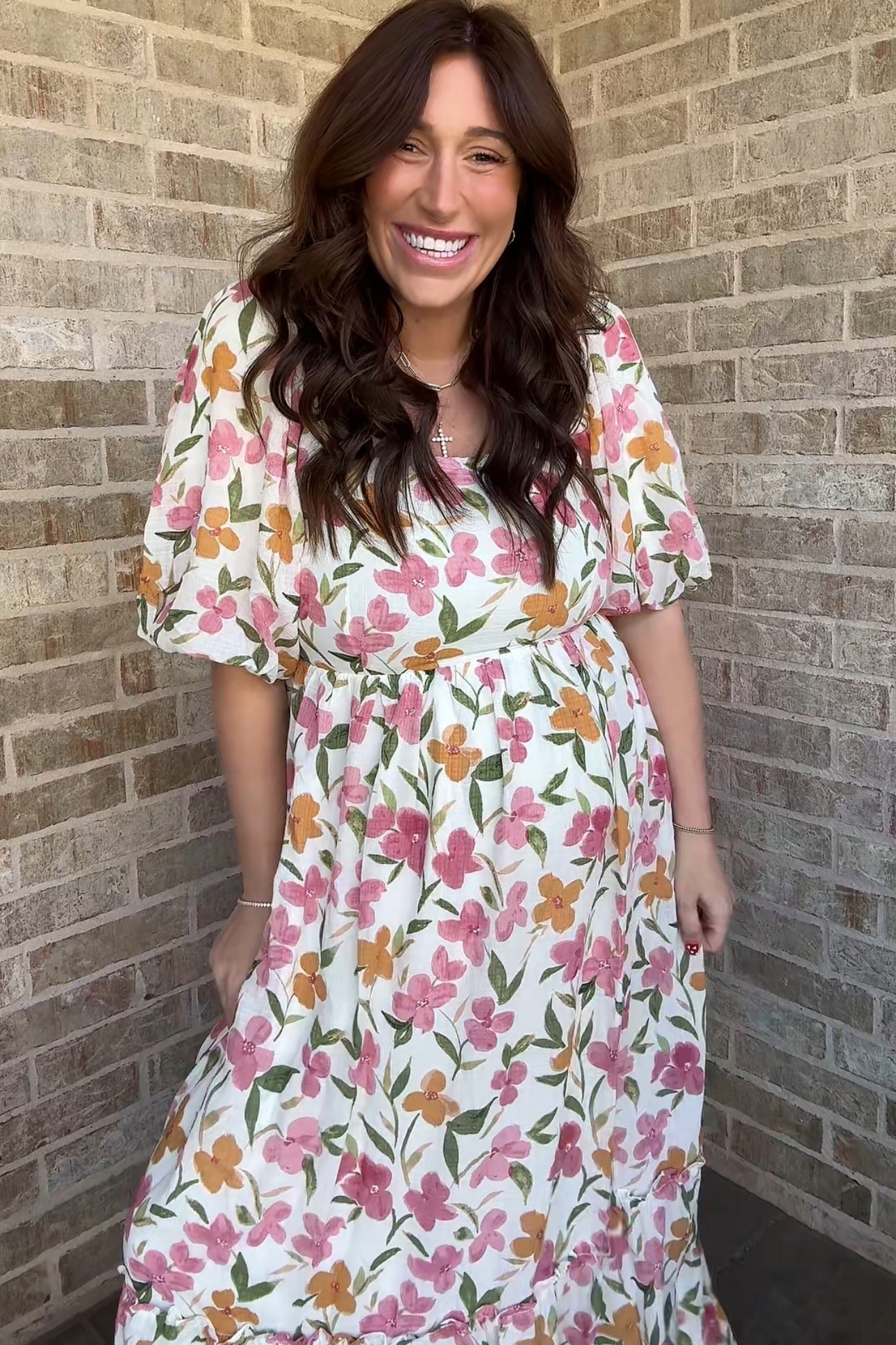 woman wearing a floral print maternity maxi dress with puff sleeves and a tiered hem, smiling in front of a brick wall