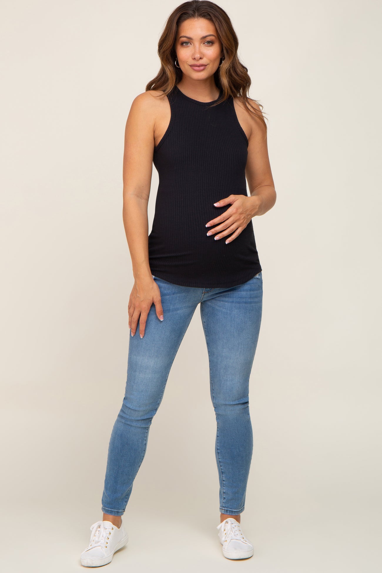 woman wearing a black ribbed maternity tank top and holding her baby bump, paired with blue skinny jeans and white sneakers