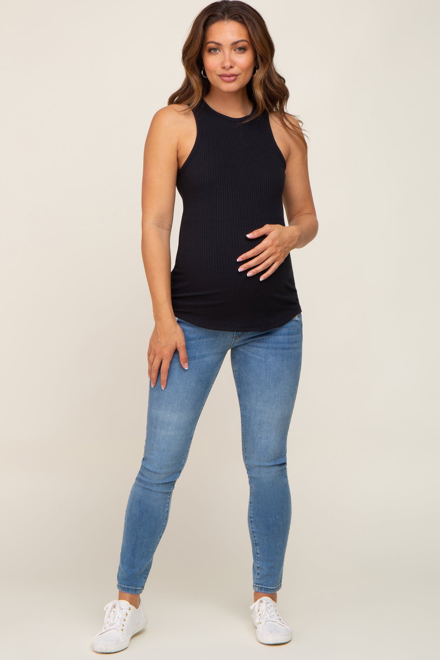 woman wearing a black ribbed maternity tank top and holding her baby bump, paired with blue skinny jeans and white sneakers
