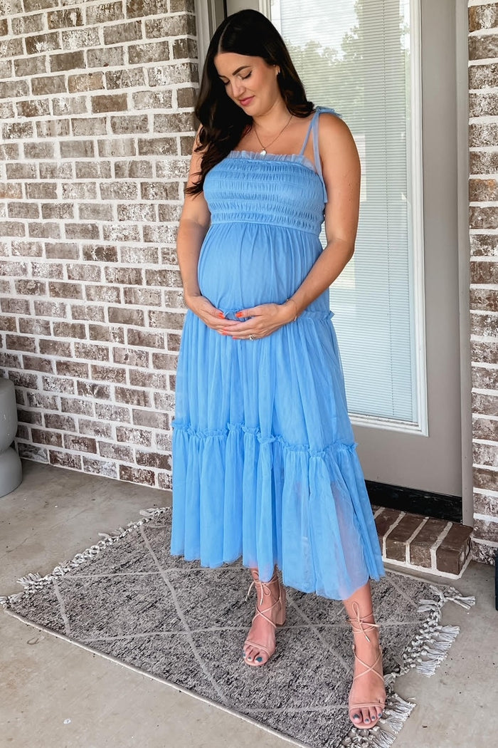 pregnant woman in a blue smocked tulle maternity dress holding her baby bump in front of a brick wall