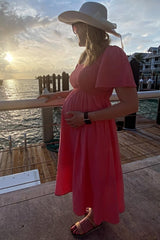 woman wearing a coral pink maternity dress with short puff sleeves and a visible baby bump by the waterfront at sunset