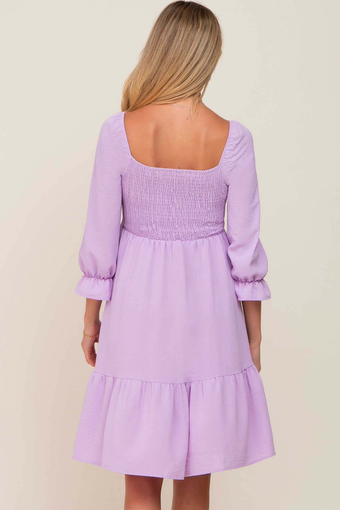 Back view of a woman wearing a lavender long sleeve smocked mini dress with a ruffled hem.