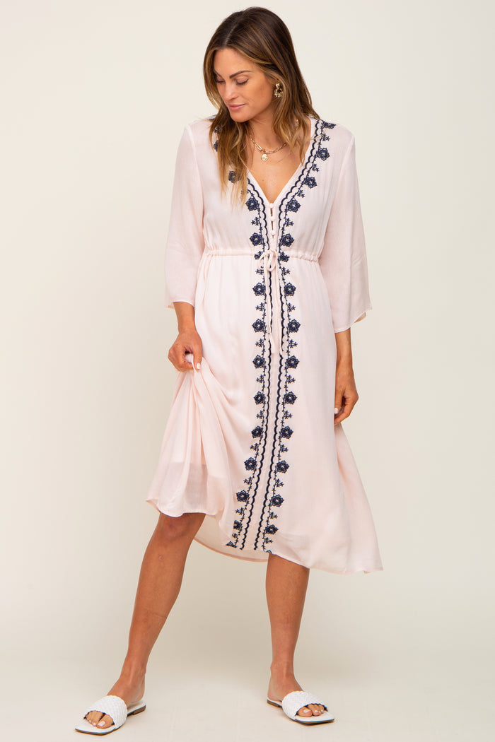 woman wearing a light pink maxi dress with navy blue embroidery and a tie waist detail, styled with white sandals