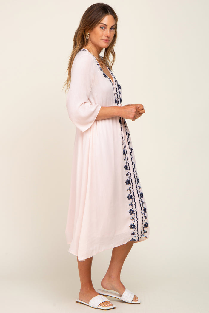 woman wearing a blush pink embroidered midi dress with bell sleeves and side slits, styled with white slide sandals