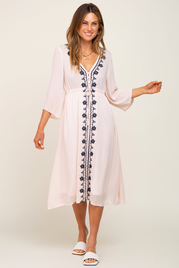 woman wearing a blush pink midi dress with navy floral embroidery and three-quarter sleeves, styled with white sandals
