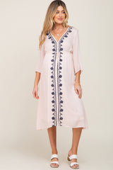 woman wearing a light pink maternity dress with navy embroidery and three-quarter sleeves, standing and smiling
