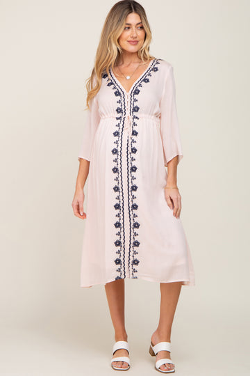woman wearing a light pink maternity dress with navy embroidery and three-quarter sleeves, standing and smiling
