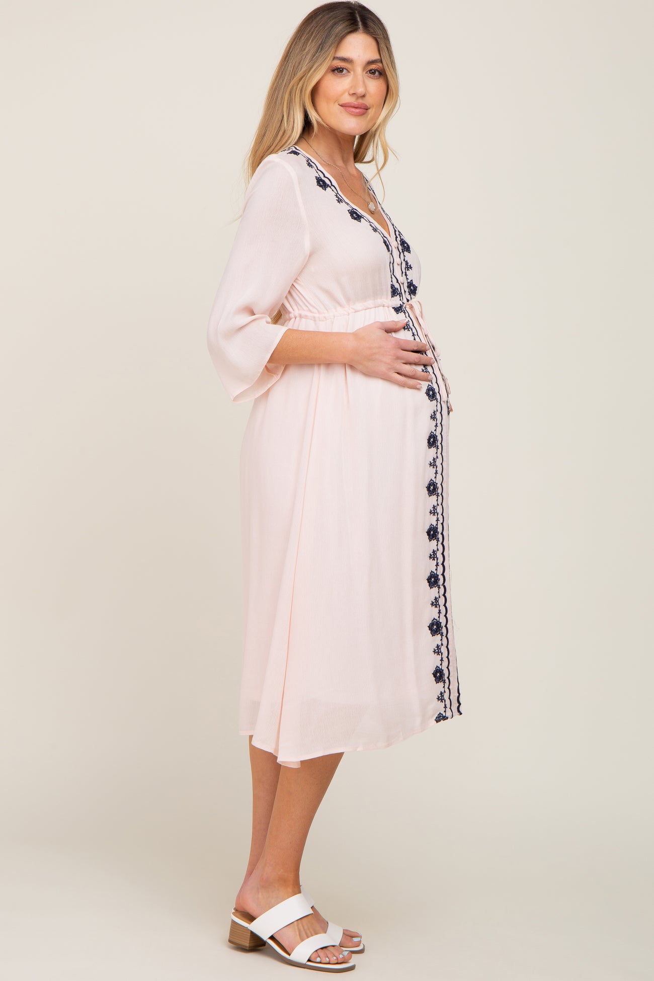 woman wearing a light pink embroidered maternity dress, cradling her baby bump while standing sideways