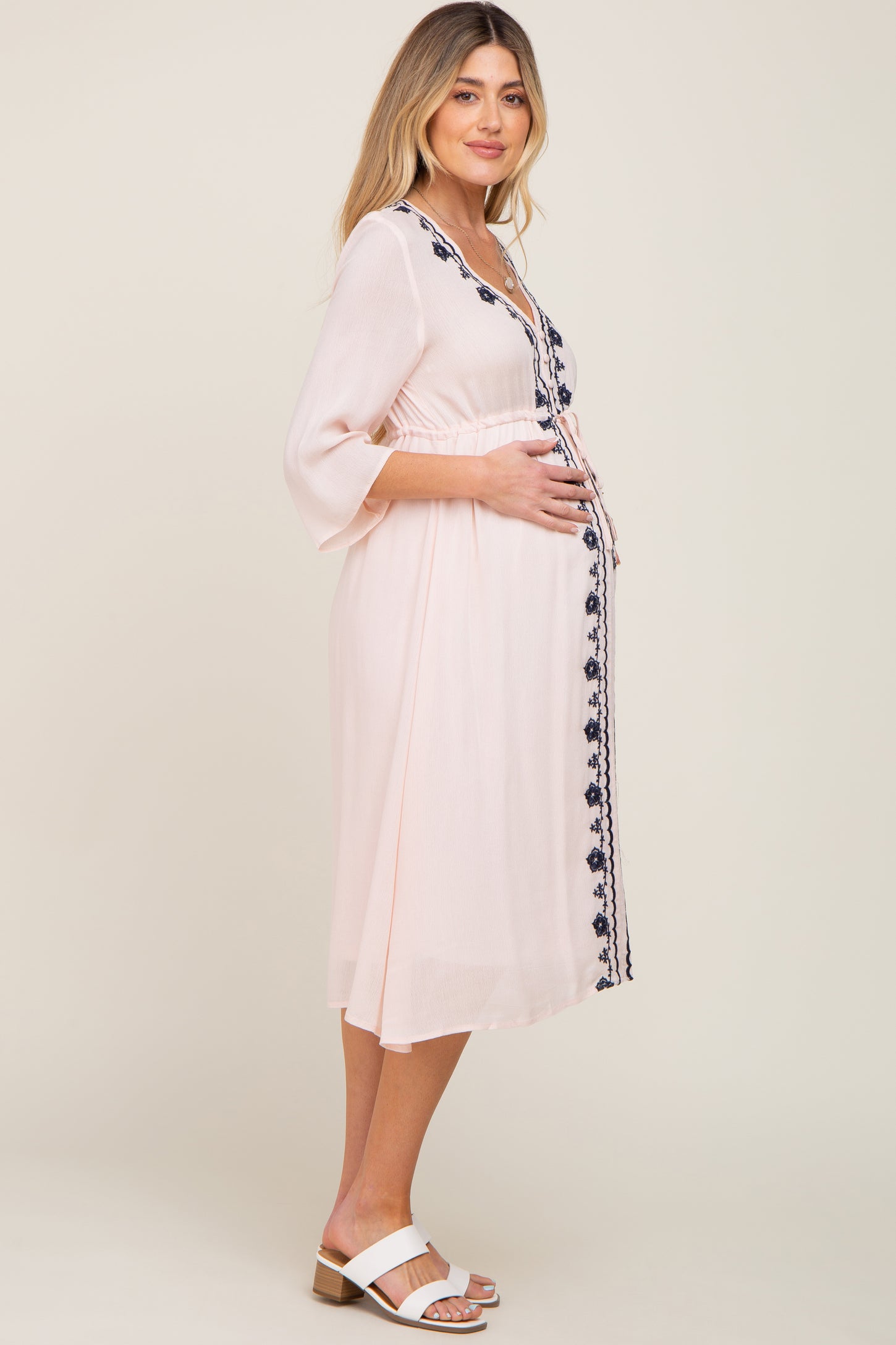 woman wearing a light pink embroidered maternity dress, cradling her baby bump while standing sideways