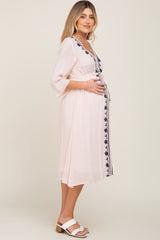 woman wearing a light pink embroidered maternity dress, cradling her baby bump while standing sideways