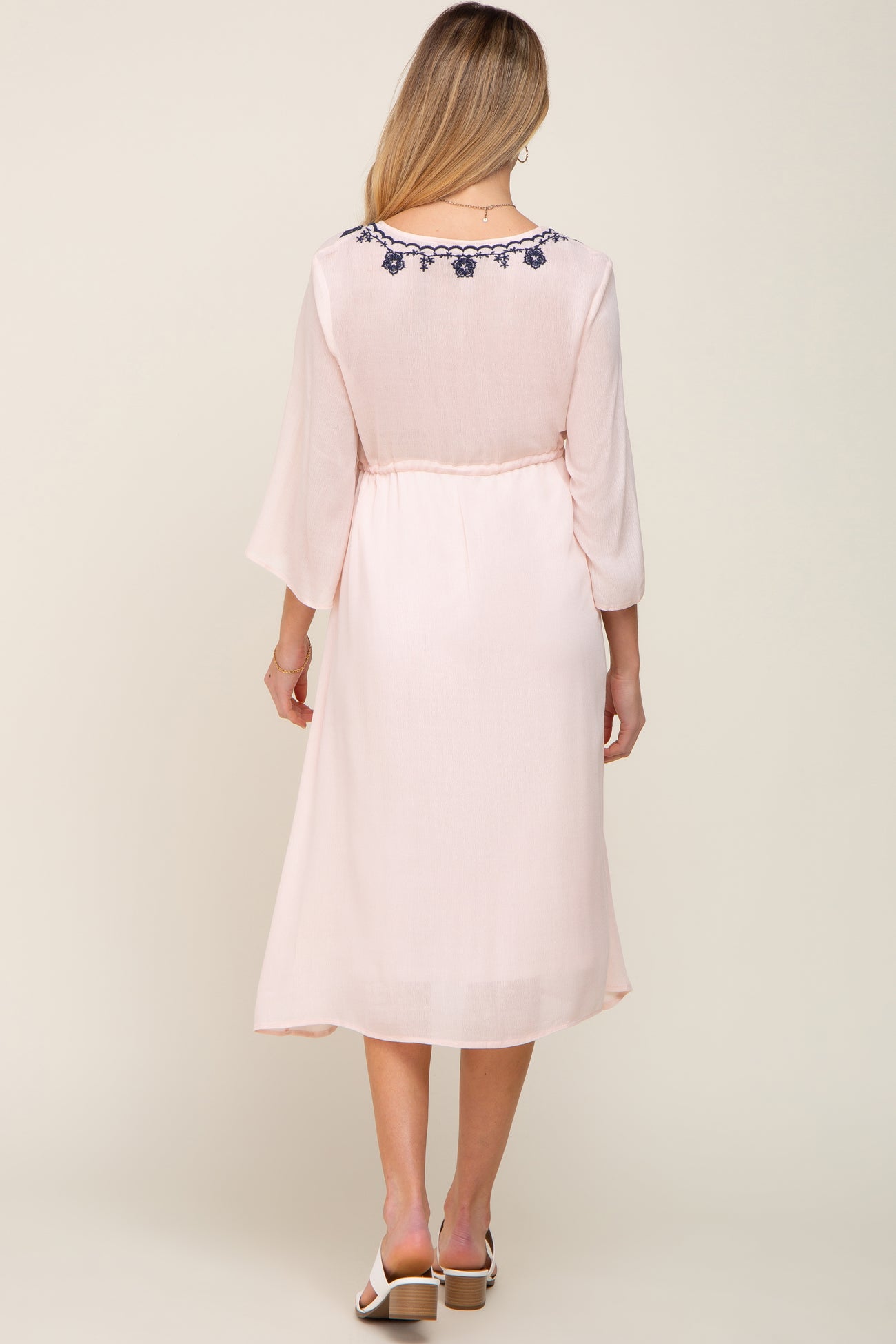 Back view of a woman wearing a light pink midi dress with navy blue floral embroidery on the neckline