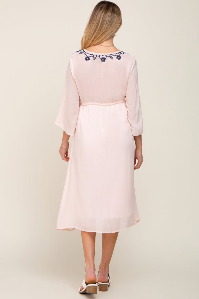 Back view of a woman wearing a light pink midi dress with navy blue floral embroidery on the neckline