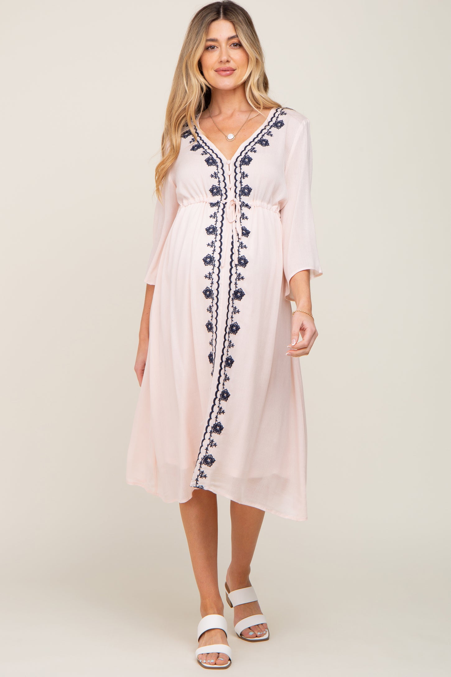 woman wearing a light pink maternity dress with navy floral embroidery and a tie waist detail
