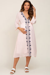 woman wearing a light pink maternity dress with navy embroidery and three-quarter sleeves, holding her baby bump