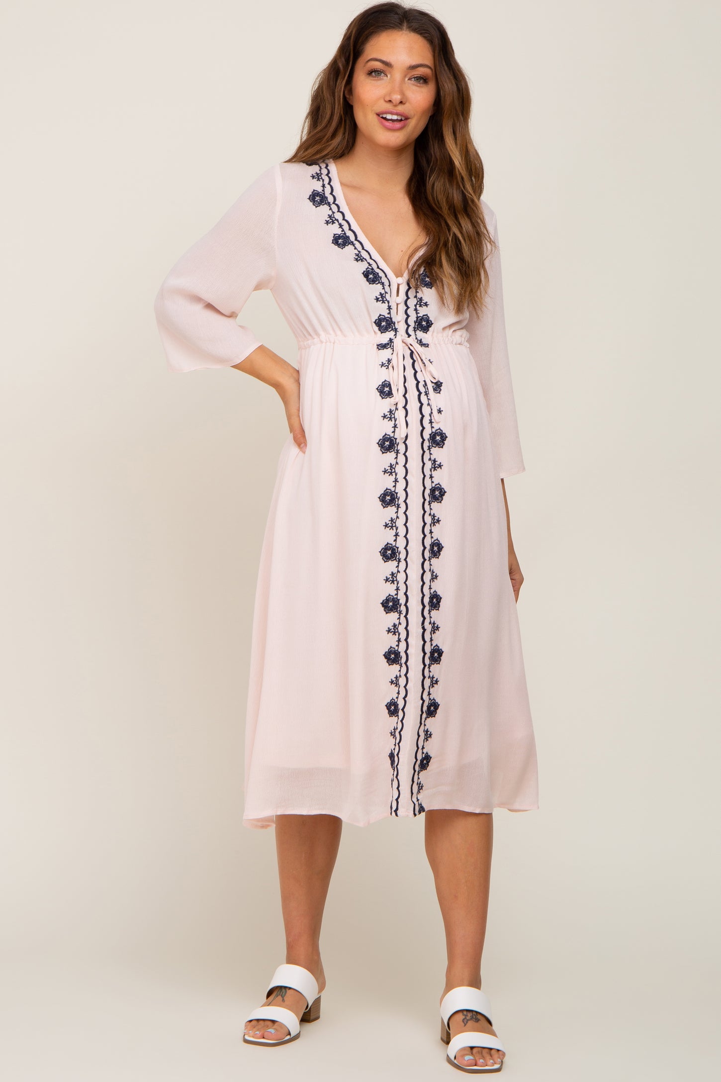 woman wearing a light pink maternity dress with navy embroidery and three-quarter sleeves, standing confidently
