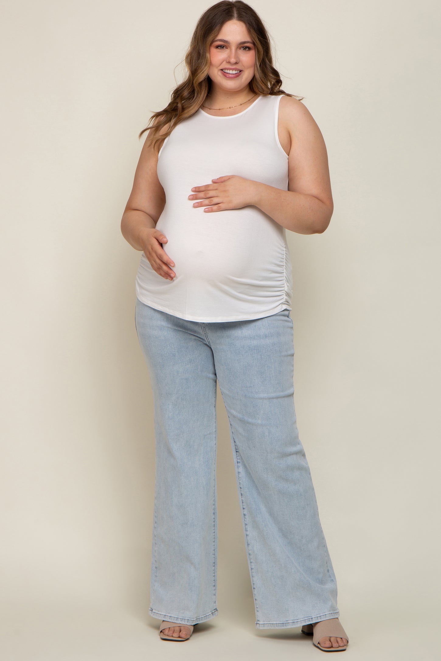 woman wearing a white sleeveless maternity top and light blue flared jeans, cradling her baby bump