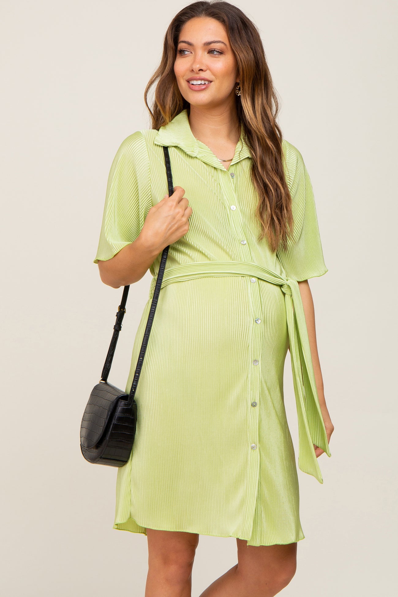 Pregnant woman in a lime green pleated maternity shirt dress with a waist tie and black crossbody bag